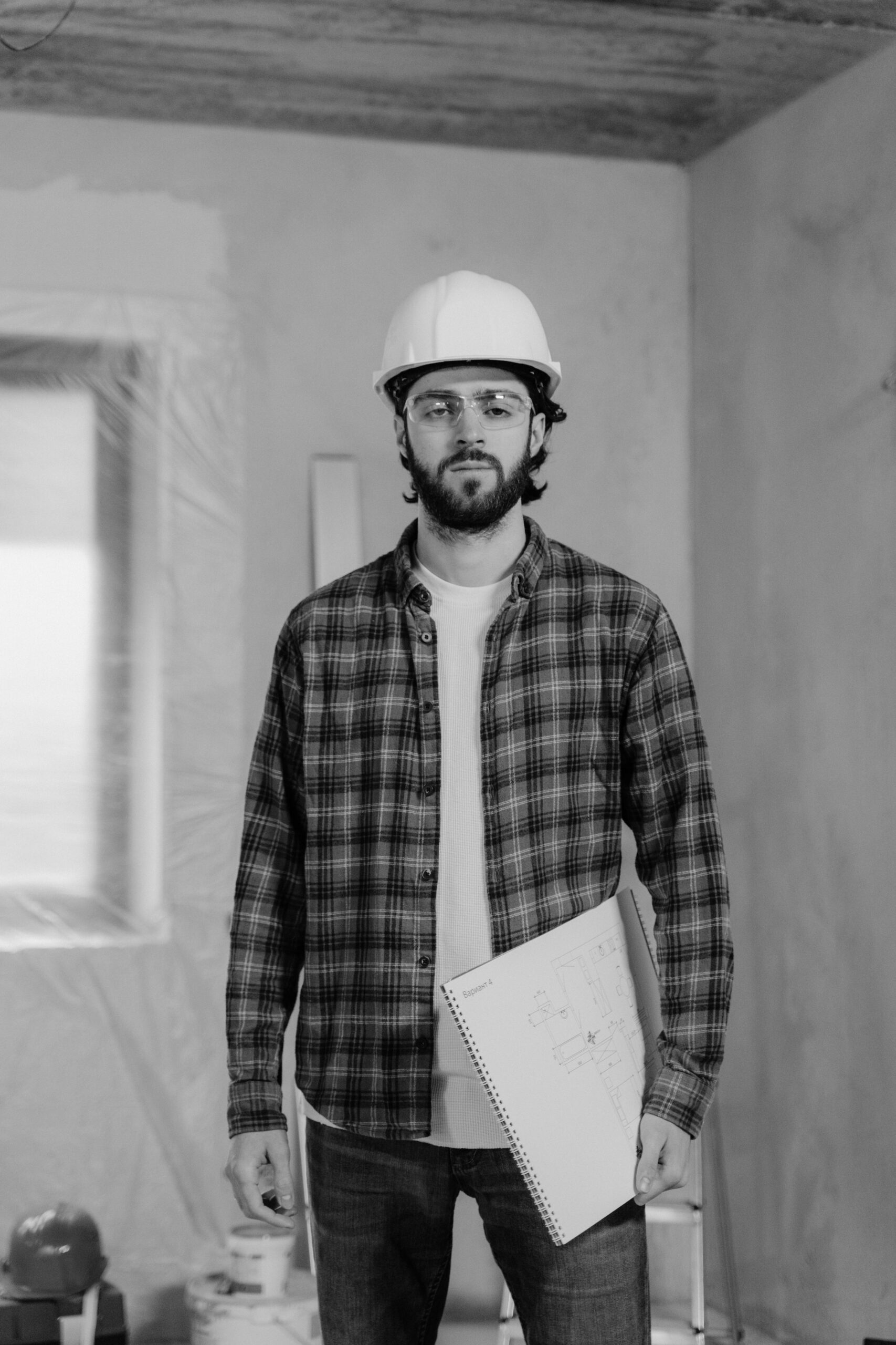 A construction worker with a hard hat and plaid shirt holding a blueprint inside a renovation site.